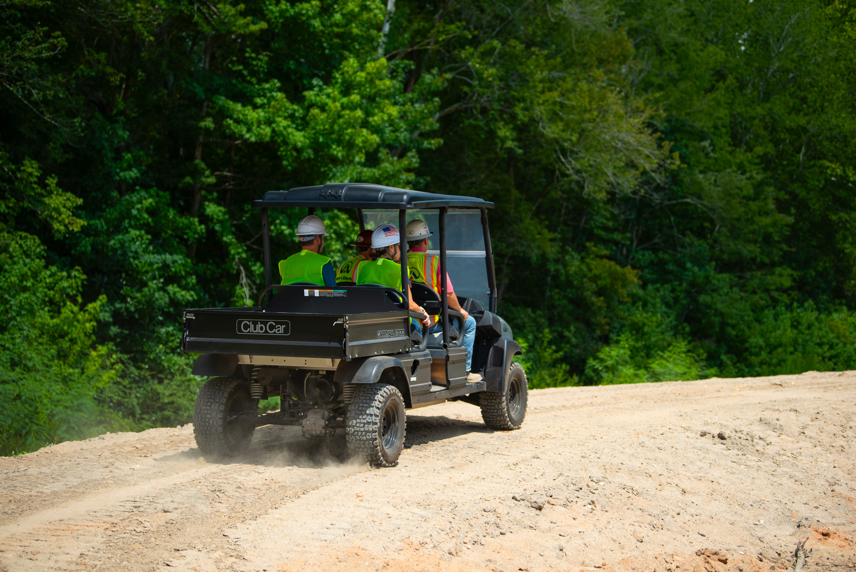 4 men in club car carry all utility vehicle wearing yellow construction safety vests driving down di…